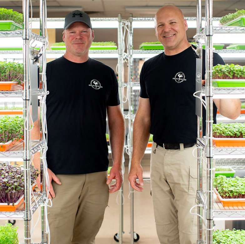 Grand Valley Micro Farms - Colorado - 06.13.24_Team_GrowRoom_Vertical Two men in black t-shirts stand in a modern indoor farm surrounded by shelves of vibrant microgreens and seedlings, showcasing sustainable agriculture practices.