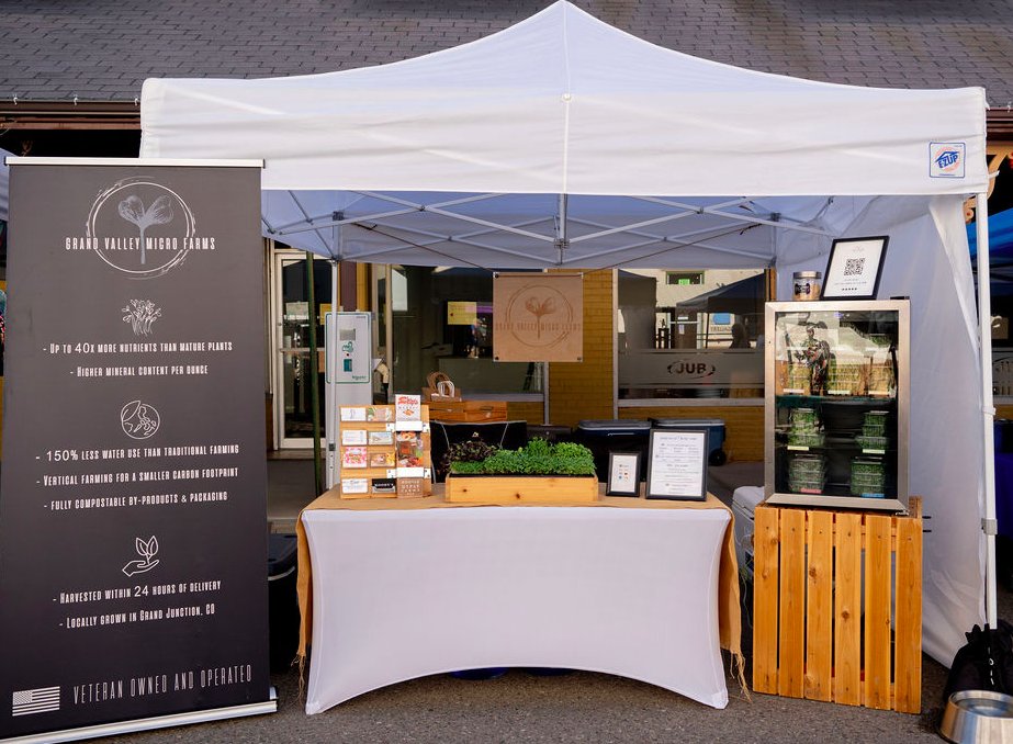 Grand Valley Micro Farms - Colorado - 06.16.24_FM_booth Grand Valley Micro Farms booth at a market, showcasing sustainable farming practices with a black banner highlighting nutrient-rich microgreens, water-efficient vertical farming, and compostable packaging. The display features fresh produce, informational materials, and a compact refrigerator with microgreens, emphasizing local and veteran-owned operations.